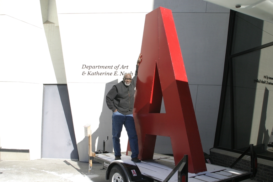 Clarence Morgan standing in front of Regis Center next to a 10 ft tall red metal letter A sculpture