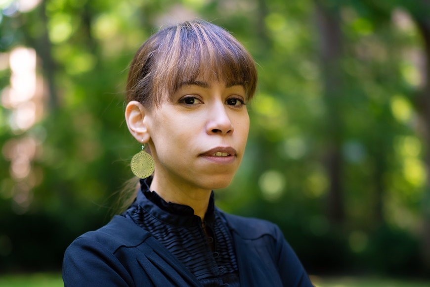 Head and shoulders of person with medium brown hair with bangs and pulled back and light brown skin, wearing dark blue top in front of green trees