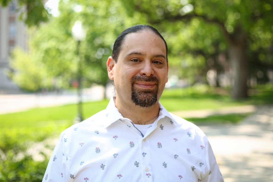 Jimmy Patiño, a man with black hair and a black moustache and goatee, wearing a white button-up shirt, stands outside on a sunny path