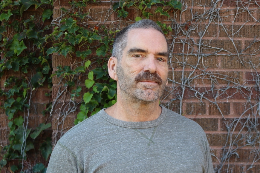 Kevin Murphy looking into the camera with a serious expression in front of a brick building with vines