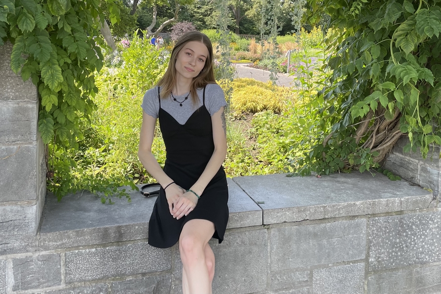 skinny young woman with pale skin and shoulder length brown hair sitting outside on a gray stone ledge in front of green fields. Wears a black knee-length dress over a gray t-shirt and black-beaded necklace. Wears a green beaded bracelet on right hand and a black ponytail hair tie on left hand