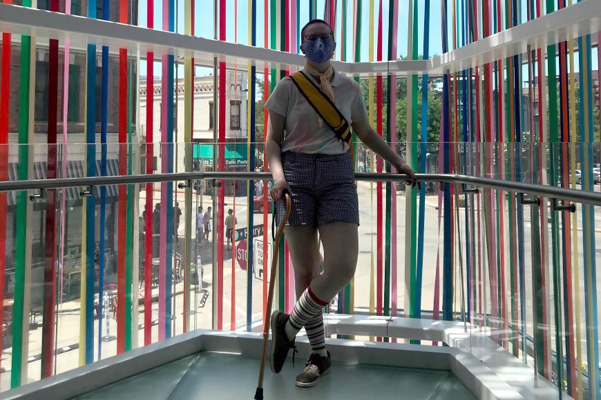 Image of person in dark shorts and light short-sleeved shirt with gold strap over chest, short hair and mask, holding cane, in window-walled space with multi-colored ribbons hanging vertically; streetscape visible beyond glass