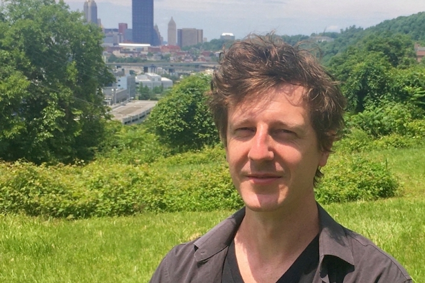Man in dark shirt on a green hill, with Pittsburgh on the skyline