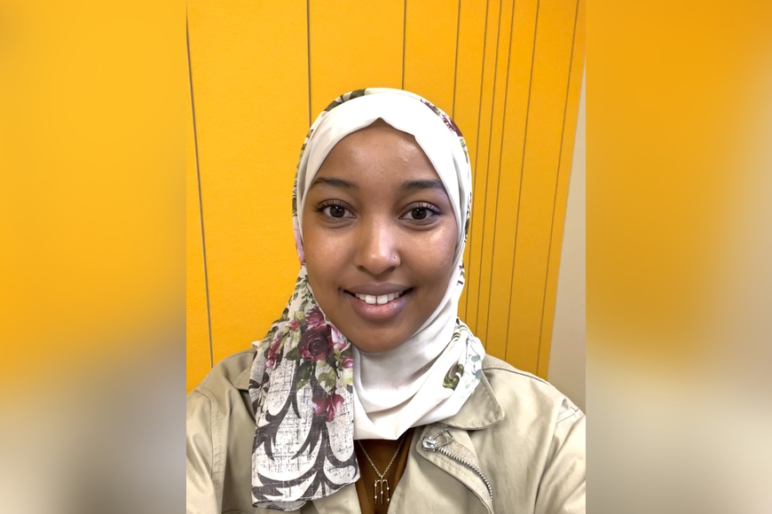 Muna Mohamed smiles at the camera wearing a patterned hijab with a yellow background