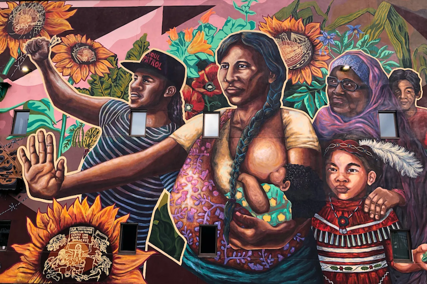 Large mural featuring a woman with a serious expression on her face. She has brown skin and a long black braid. She is nursing a baby and holding a hand out palm forward. She is surrounded by other Black, Indigenous, and People of Color. Around them are sunflowers and other plants.