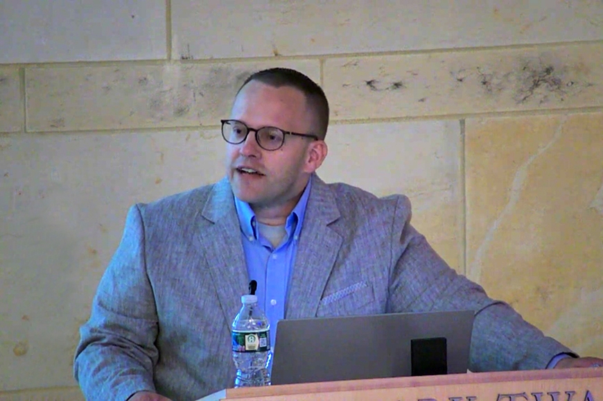 Head and upper torso of person with brown crewcut, faint beard, and light skin, talking and wearing glasses and light blue collared shirt and suitcoat; yellow and white stone wall behind and lectern, water bottle, and laptop in front