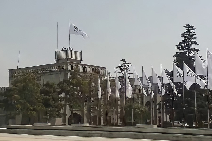 Afghanistan presidential palace with the Taliban's white flag with black symbols flying from its roof. A row of white Taliban flags on flagpoles are in the foreground