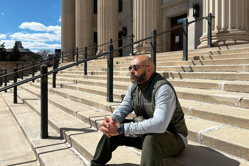 A man with a beard and sunglasses sits contemplatively on the steps in front of Northrop Auditorium