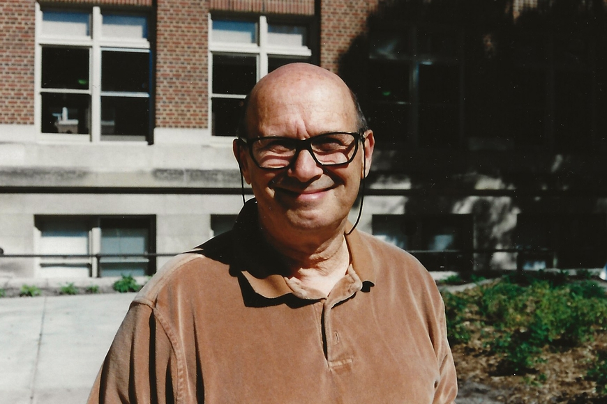 Photo of person with bald head, glasses, pale skin, wearing brown shirt, standing in front of brick building