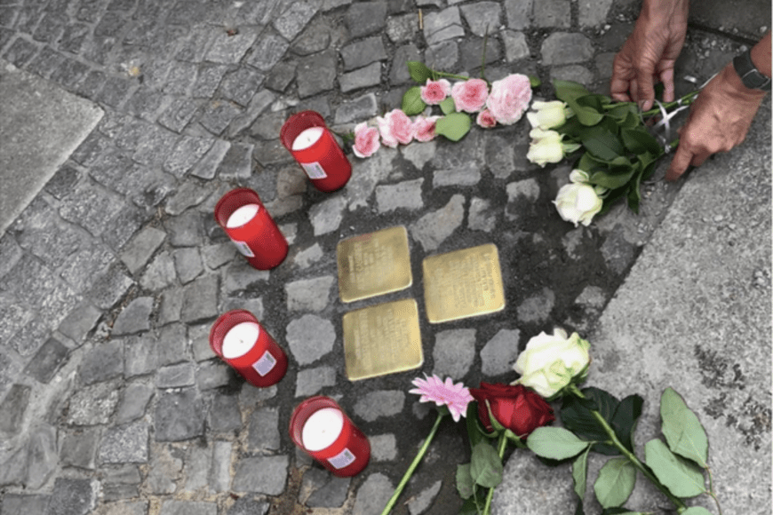 an image of several Stoplersteine, small brass plates put into the ground outside of the homes of Holocaust victims, surrouned by 4 red candles and flowers. 