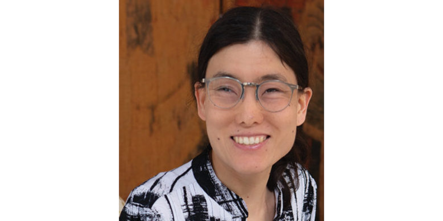 Person with dark hair tied behind their head wearing circularly framed silver glasses smiling. The person is wearing a black and white striped design shirt. 