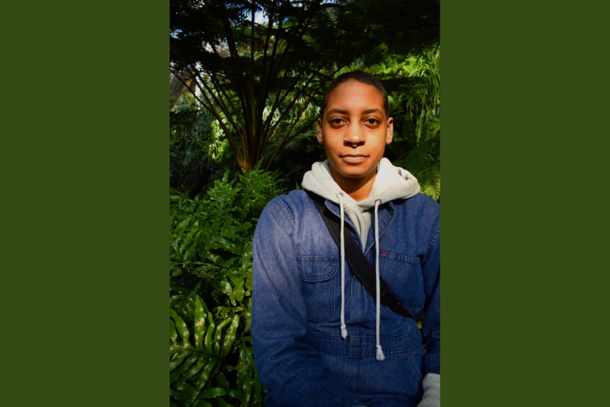 Image of Moriah Shumpert, wearing a hoodies and jean jacket, standing in front of a lush forest