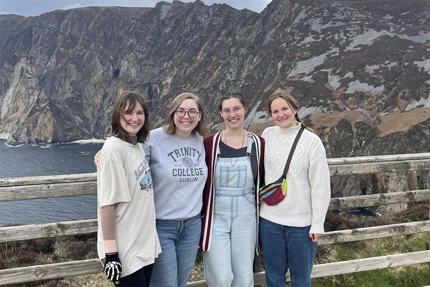 Four young women - Cece Roth, Julianna Bruemmer, Claire Voskamp, Stella Mehlhoff - stand in front of large hills and a body of water