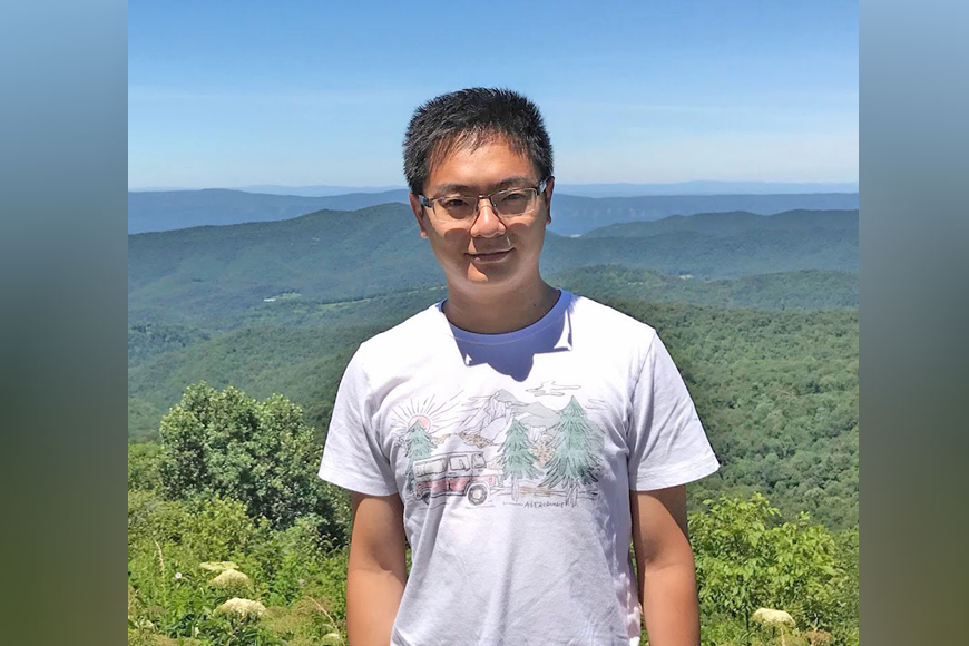 Asian man with black hair, white T-shirt, and glasses, with large expanse of forest below and behind him.