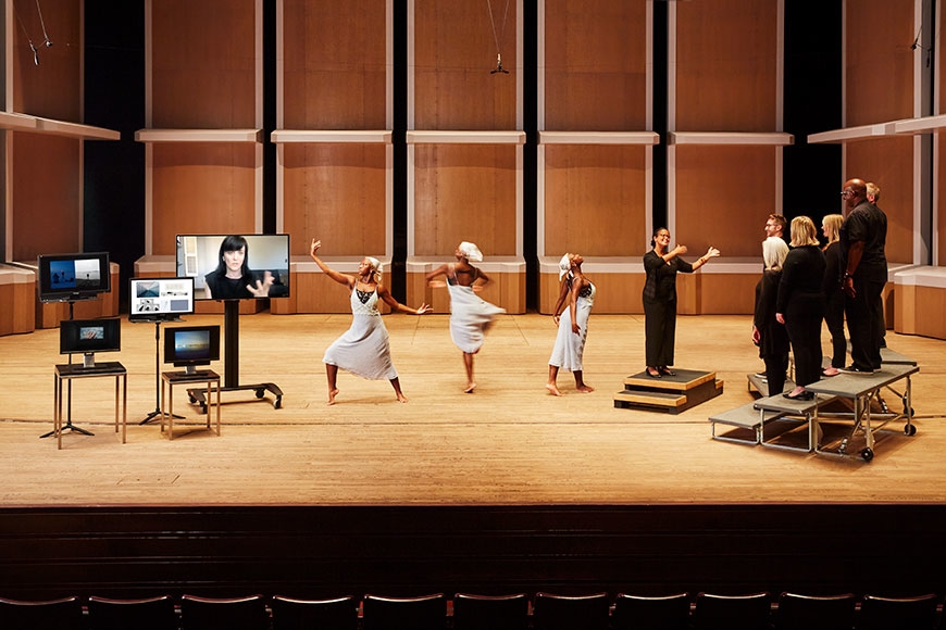 Image of Ted Mann stage, featuring digital monitors, a dancer, a choral director, and a small choir.