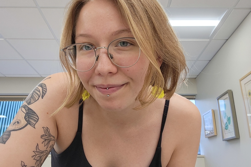 Girl with white skin, shoulder length blond hair, a black floral tattoo on left arm, lip piercings, and silver glasses, wearing a black tank top and yellow feather earrings. Girl is smiling in a classroom with white walls, three pictures on the right, white ceiling, and window 