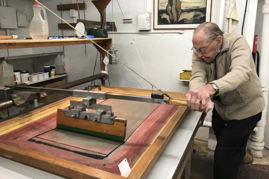 An elderly man operates the squeegee on a screenprinting press