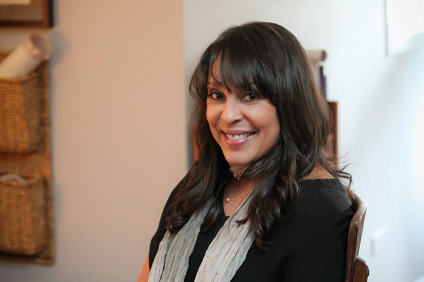 Head and shoulders of person with long dark hair and bangs and light brown skin, smiling and wearing dark top with grey scarf around neck