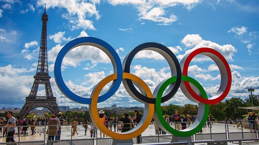 An Olympic rings sculpture with the Eiffel Towel in the background with a blue sky and clouds behind them