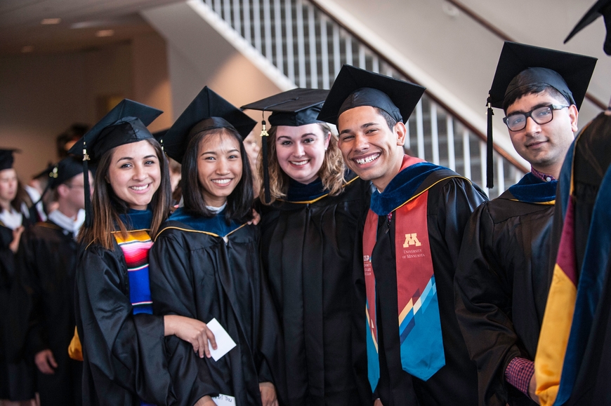 five diverse and smiling students stand together in their graduation regalia, 