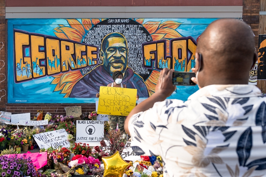 Photo of the George Floyd Memorial in Minneapolis. A person wearing a white mask is holding a yellow sign that says "George Floyd deserved to live"