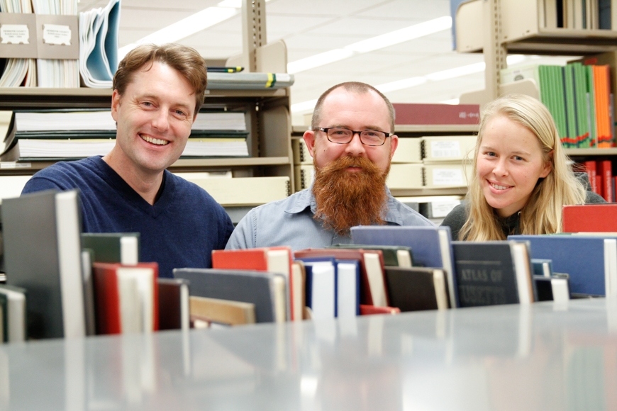 Three people pose together in the Borchert Map Library. They are looking over a bookshelf at the camera and smiling.