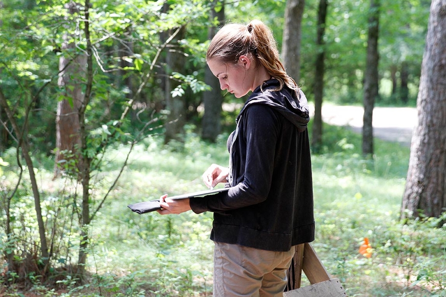 Young woman outdoors in a wooded area. She is holding a notebook and writing in it.