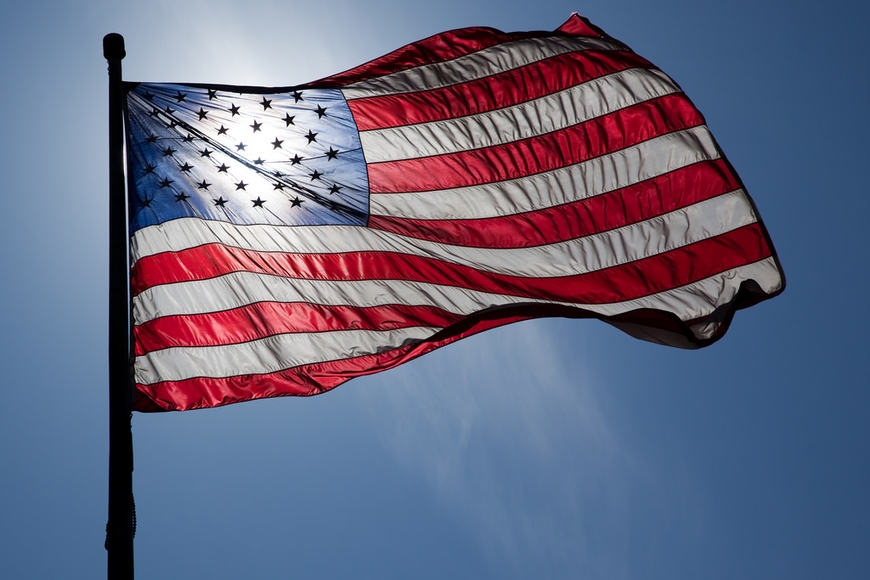 An American flag on a flagpole, blowing in the wind and blacklit by bright blue sky and sunshine