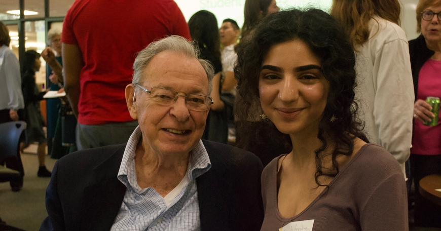 older caucasian man in a blue blazer sits next to young brown skinned woman with curly hair