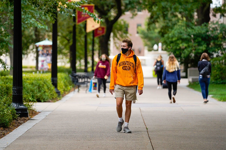 A young man walking on Northrop Mall wearing a University of Minnesota hoodie and a face mask.