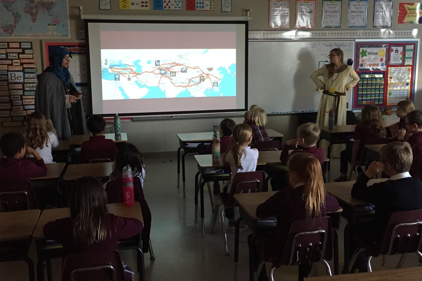 Graduate students, dressed in costumes from medieval western Europe and northern Africa, discuss the silk road with a class of elementary students seated in desks. There is a depiction of the silk road projected on the front board.