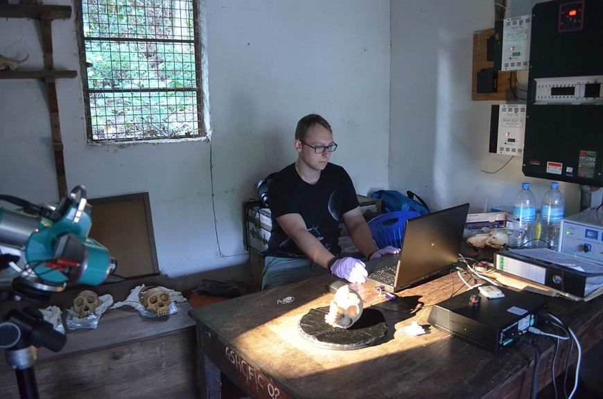 PhD Student Jason Massey sitting at a table in Tanzania. He is wearing latex gloves. Several skull models are on the desk and on tables around him.