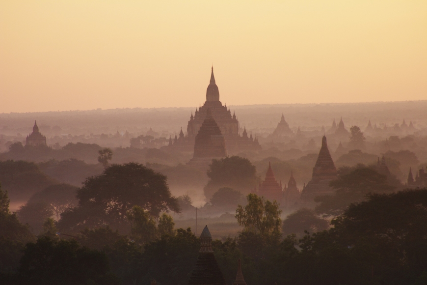 An image of a lanscape with the dark outline of trees in the foreground and shadows of large temples behind them. The sky is a pale yellow and there's a smoky fog over the whole scene.