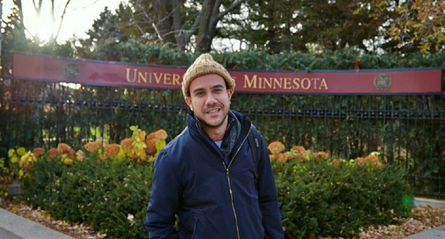 Nico Ramos Flores standing outside, with University of Minnesota sign, trees and bushes in background