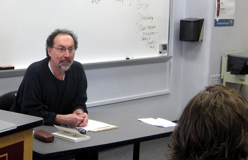 Professor Emeritus Geoffrey Sirc at table in classroom smiling at students (visible is one student's head from the back)