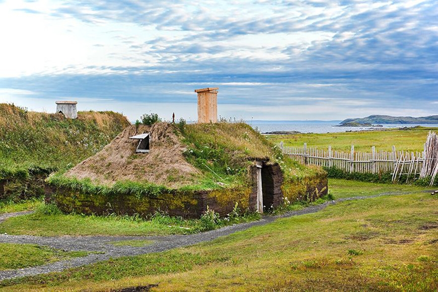 Photo of reconstructed Norse settlement called L’Anse aux Meadows in Newfoundland, Canada. The low building has a thatched roof and a chimney.