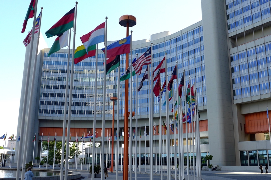Many different country flags fly on flagpoles in front of a large, glass-windowed building