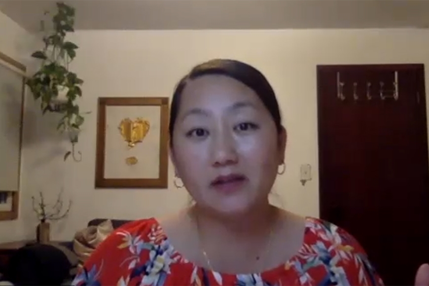 Screenshot of author Kao Kalia Yang doing Zoom call from home, head and shoulders, with white walls behind, a hanging green plant, one gold framed artwork, and dark brown door