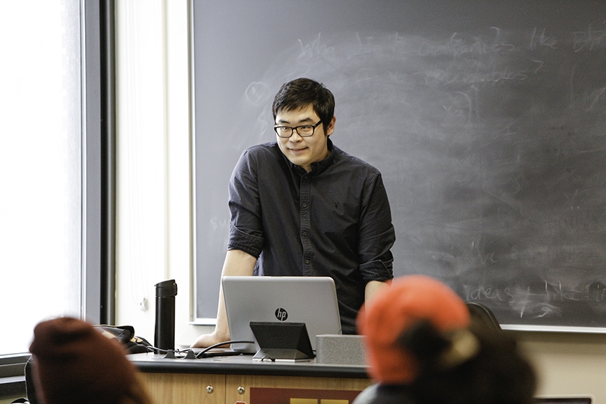 PhD candidate Youngbin Hyeon teaching in front of blackboard with laptop in front and the backs of two students' heads in foreground