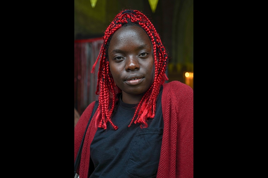 Photographic portrait of a young Black woman with bright red braids looks serenely at the camera as she tilts her head slightly to the side.