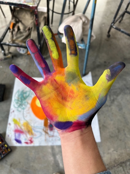 A close up image of the artist’s palm, covered in a spectrum of colored pastels, loosely resembling a heat signature. In the background behind his hand are art materials and his belongings strewn across a classroom floor.
