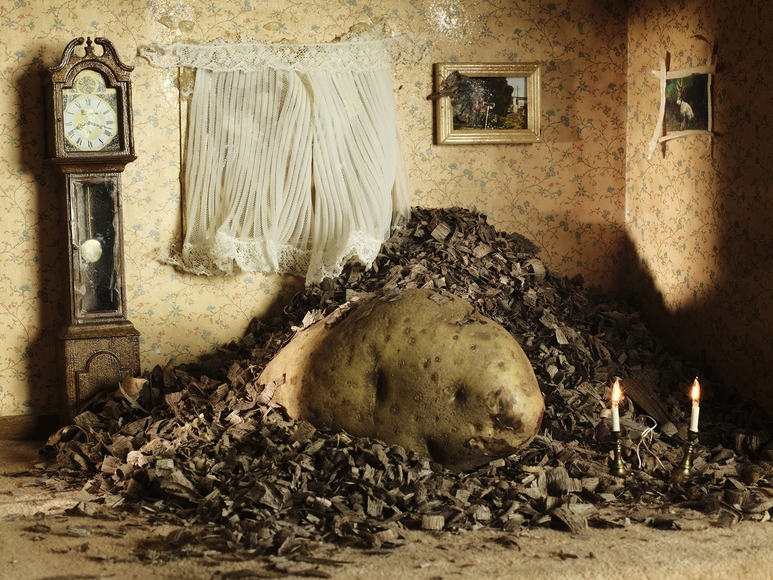 A potato sits in a pile of wood shavings appearing enormous inside a miniature bedroom complete with small electric candles on the floor and a grandfather clock against the wall.