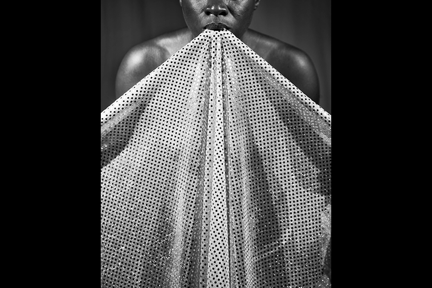 Photographic self-portrait of a Black woman from the nose down with bare shoulders who holds a sparkly polka dot fabric draped from her mouth to create a triangular shape that covers her body from view.