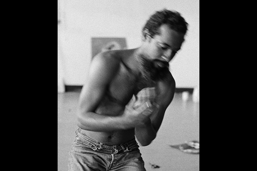 Photographic portrait of African American conceptual artist David Hammons seen shirtless and bent over holding his hands together in his Los Angeles studio.