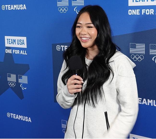 A young Hmong woman with light skin and long black hair and wearing a white sweatshirt in front of a blue press conference wall holds a microphone and smiles
