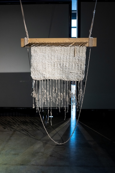 A weaving created from the torn strips of a cotton bed sheet is hanging from the wooden peg loom it was woven on. 