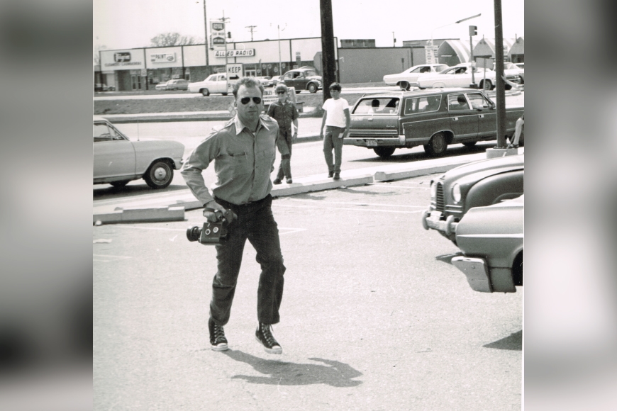 Luther Gerlach running in a parking lot and holding a camera. Cars from the time period can be seen in the background.