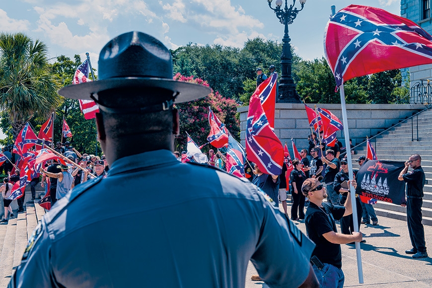 Photographic image of the back of a Black state trooper overlooking a white supremacist group protesting in front of a government building in South Carolina. White supremacists actively wave the confederate flag and hold up pro Ku Klux Klan signs.