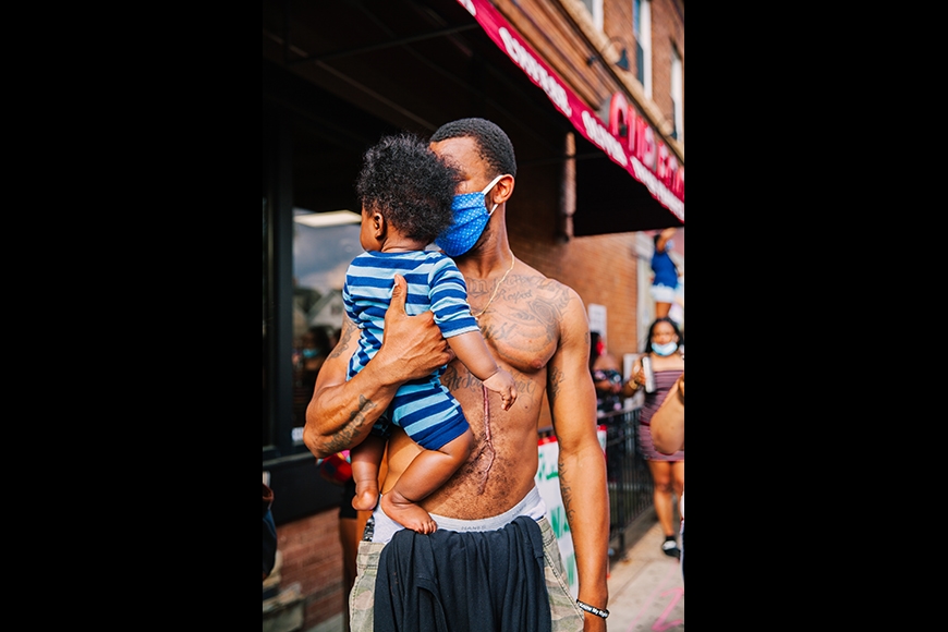 Portrait of a shirtless and masked Black father, with a large scar that runs down the length of his stomach, holds his son during a gathering at George Floyd Square the Minneapolis during the summer of 2020.