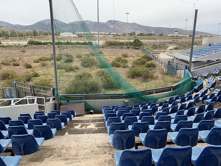 Rows of blue stadium seats facing an area with grass and bushes where a field used to be